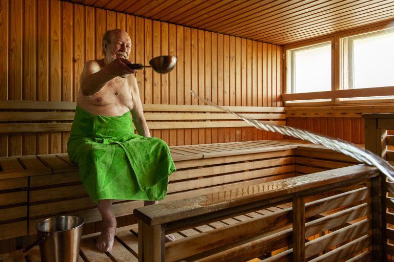 Iäkäs henkilö heittää löylyvettä saunassa pyyhe yllään.
